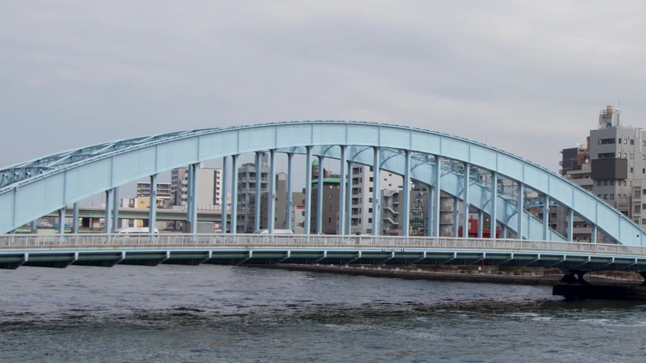Static wide shot of a blue arched bridge over flowing water under overcast skies