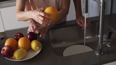 Una-Pareja-Amorosa-Irreconocible-Lava-Frutas-Frescas-En-El-Fregadero-Antes-De-Comer-Por-La-Mañana-En-La-Cocina-Moderna.-Hombre-En-Jeans-Acariciando-Suavemente-La-Espalda-De-Su-Cariñosa-Esposa