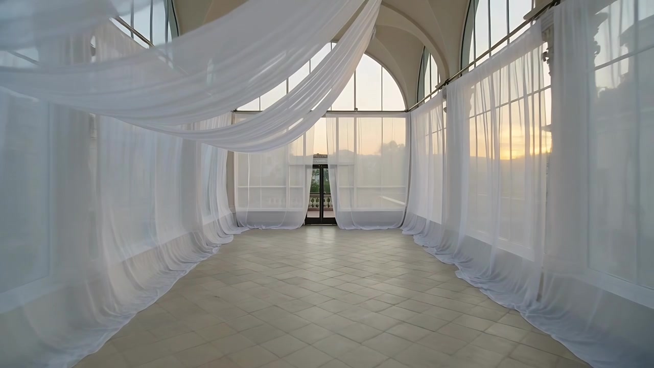 Drappeggio leggero e ondeggiante lungo un corridoio ad arco, mosso da una brezza proveniente da destra, che guida lo sguardo verso una porta in fondo.