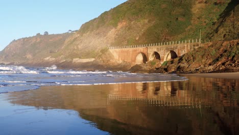 olas rodando hacia la playa con la montaña y el viejo puente del tren en el fondo