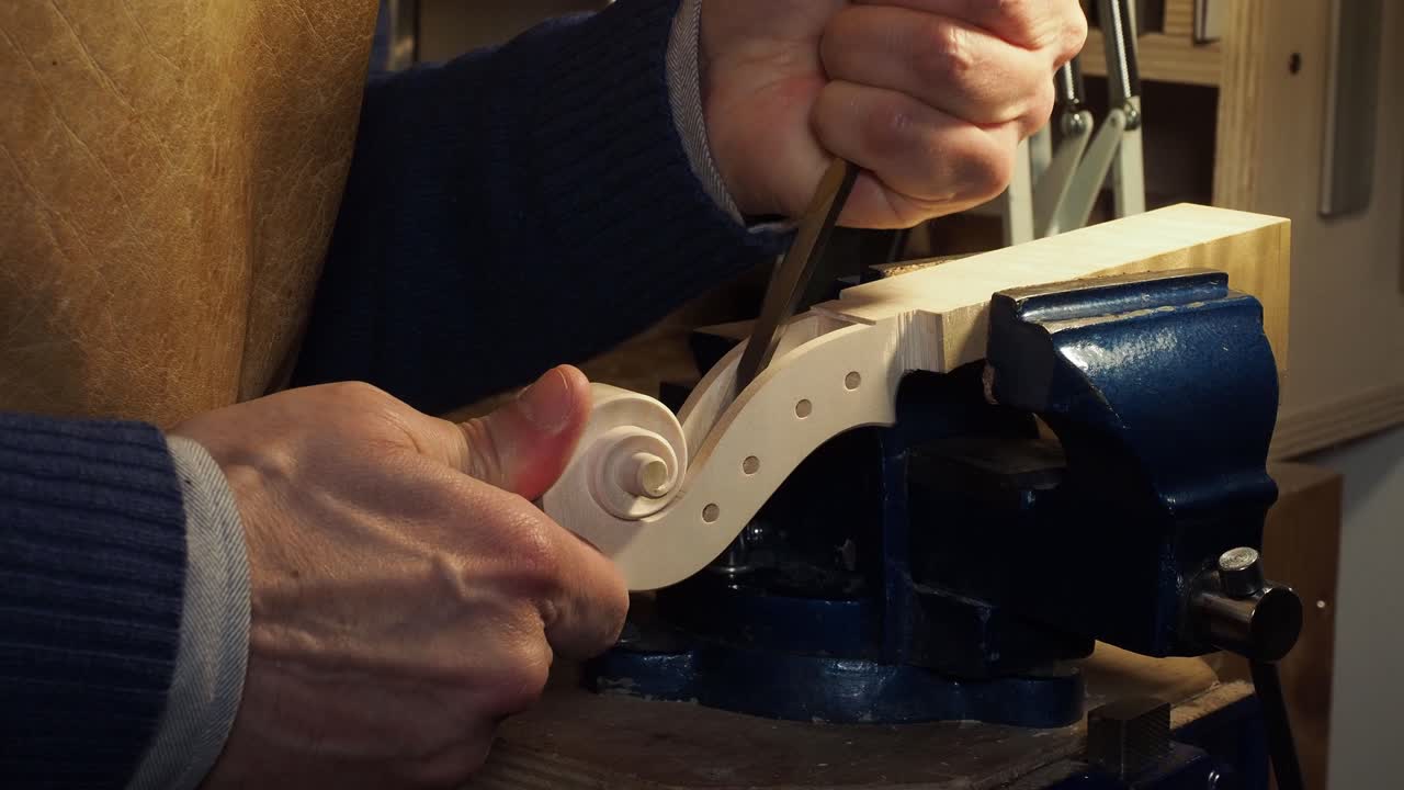 Artist using a chisel and mallet to carefully carve classic violin scroll in his workshop, the wood piece is securely clamped in a bench vise to keep it stable during process