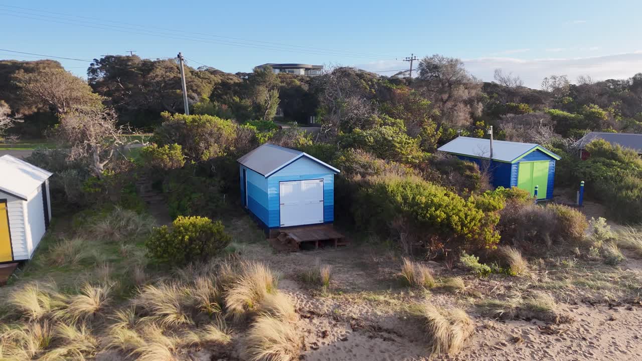 Drone glides above vibrant beach huts, sandy beach, and coastal vegetation in soft daylight
