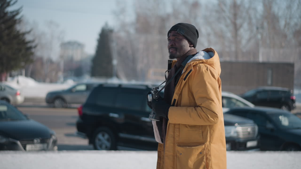 young dark man press photographer walking street holding camera gear with gloved hands while scanning snowy bustling urban scene with bare trees lined parked cars under clear blue winter sky
