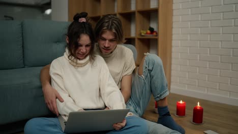 young adult couple using laptop computer at home sitting on floor around candles