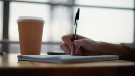 close-up of hand writing in notebook on wooden table with beverage cup and blurred background showcasing structure, creating a cozy and focused work or study environment