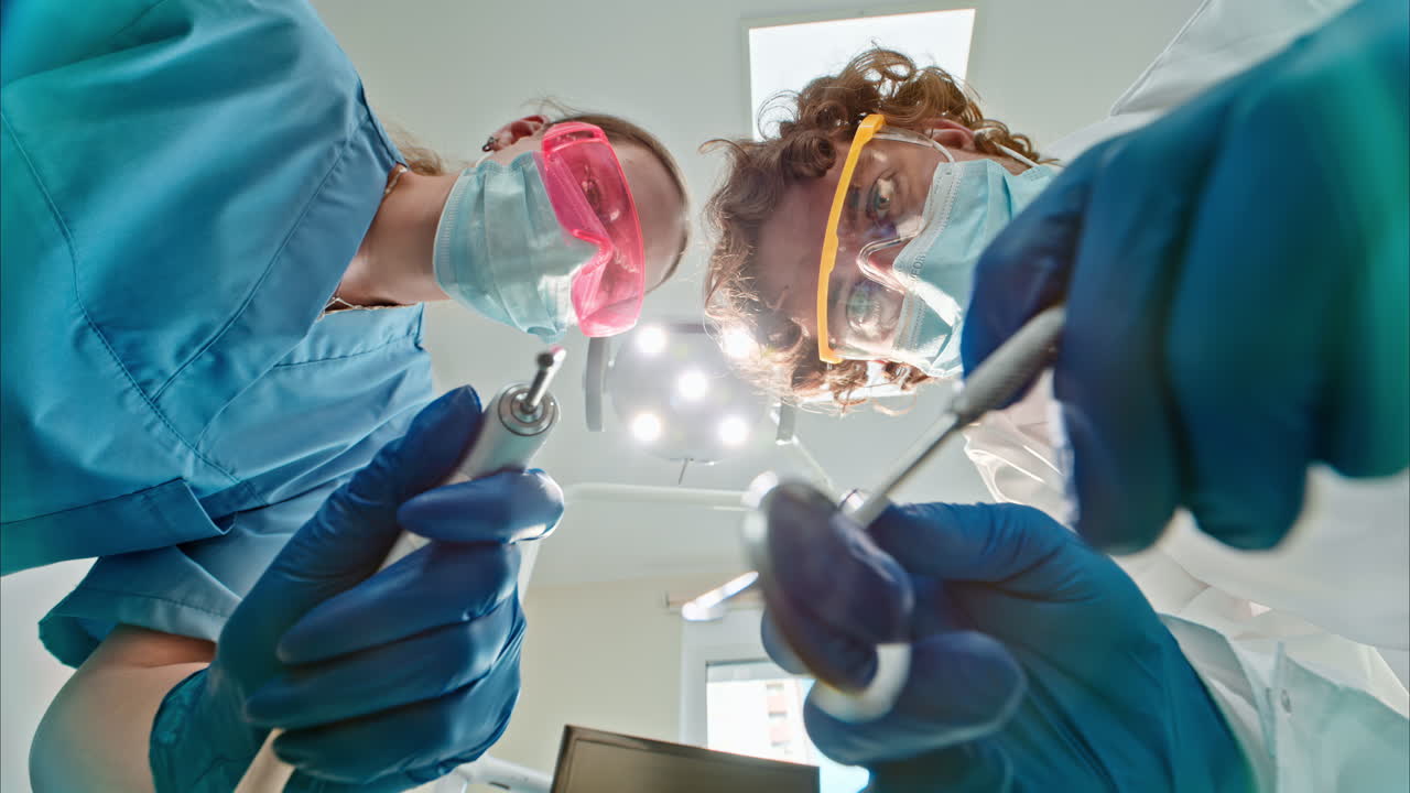 Low angle view of two dentists wearing protective masks, goggles, and gloves, holding dental instruments under bright procedural lights