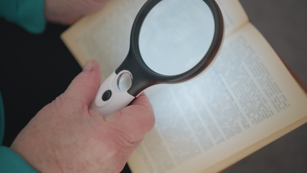 Close-up of elderly person holding magnifying glass and reading book. Focus on magnifying glass illuminating text, highlighting effort to read fine print, soft lighting, and relaxing indoor atmosphere