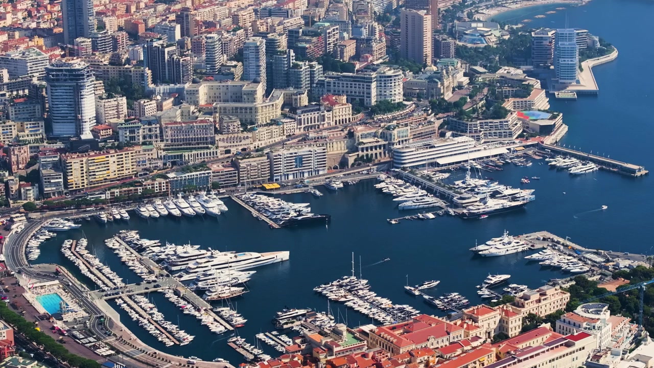 Il porto di Monaco espone una collezione di yacht di lusso ormeggiati lungo il lungomare. Edifici a molti piani incorniciano la costa, mettendo in mostra lo skyline vibrante della città durante il giorno.