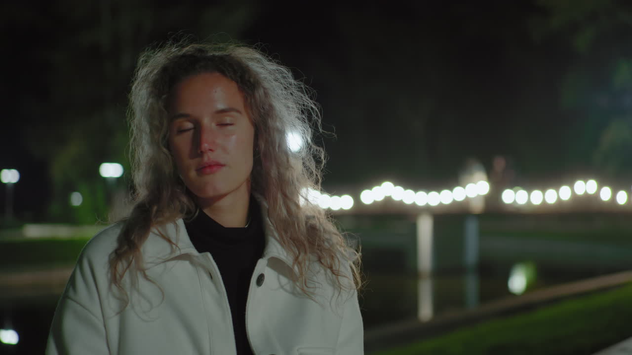 Reflective white woman with curly hair seated outdoors at night, gentle breeze swaying hair, blurred reflection behind capturing quiet mood, soft glow from illuminated background
