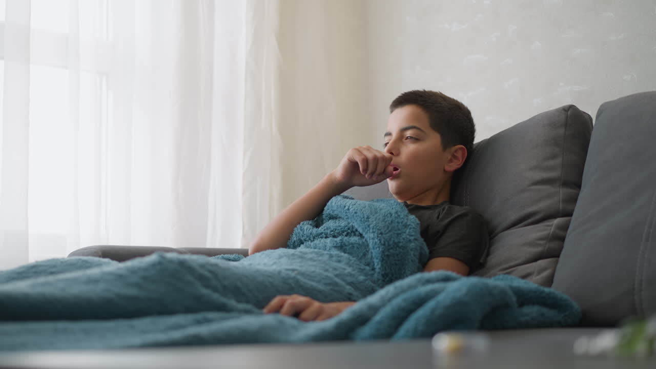 Bambino con tosse influenzale sdraiato sul divano coperto da una spessa coperta, esprime stanchezza mentre si tocca la testa e tossisce in una stanza leggermente illuminata vicino alla finestra con una tenda bianca trasparente