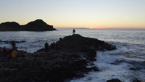 aerial track over giant’s causeway as tourists pose at sunset by crashing waves
