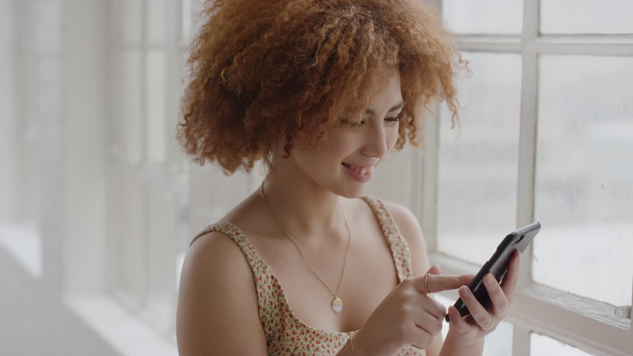 ritratto di una giovane donna di razza mista che usa uno smartphone, felice di leggere i messaggi, che si diverte a mandare sms, che comunica con il telefono cellulare, che si trova vicino alla finestra