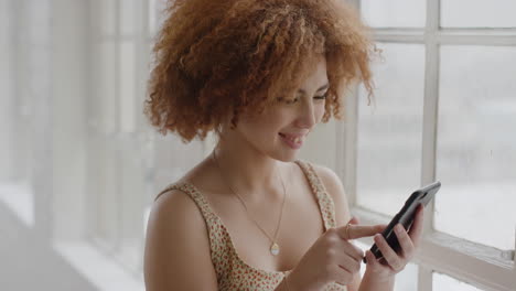 portrait of young mixed race woman using smartphone happy reading messages enjoying texting mobile phone communication standing by window