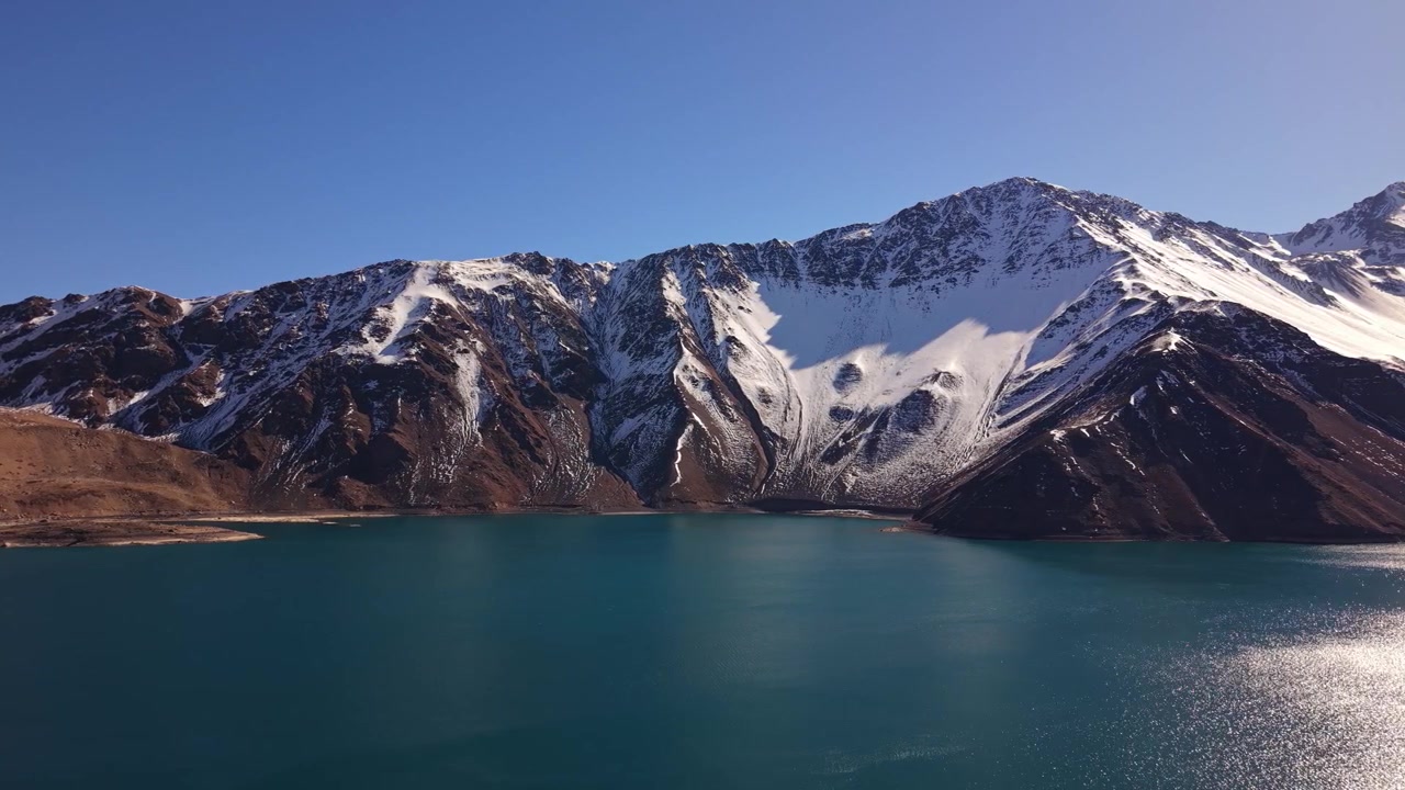 Vista panoramica di un serbatoio turchese immobile che riflette drammatiche cime andine innevate con la luce solare intensa che scintilla sulla superficie calma dell'acqua. Embalse del Yeso, Cile.