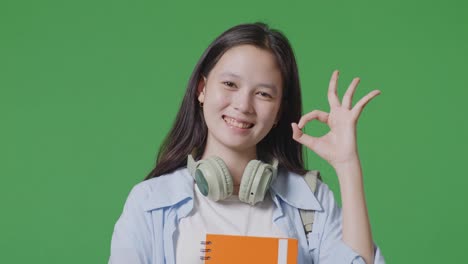 close up of asian teen girl student with a backpack and some books smiling and showing okay gesture while standing in the green screen background studio