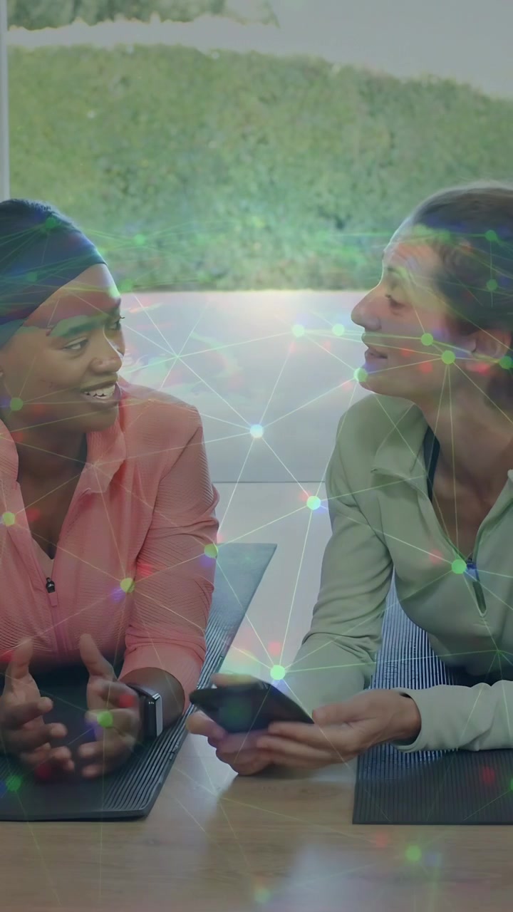 Vertical video: Right woman tapping smartphone showing workout while friend leaning, smiling