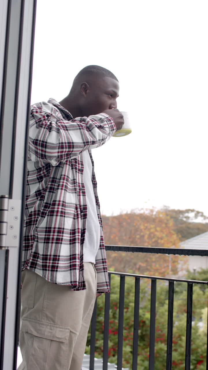 Vertical video of african american man standing on balcony and drinking coffee, slow motion