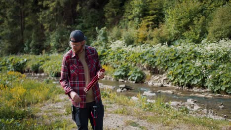 A-young-male-tourist,-blond-in-glasses-with-a-beard-in-a-red-hiking-shirt,-unpacks-the-tent.-The-guy-opens-the-frame-of-the-tent-and-tries-to-fold-it-into-a-single-structure-against-the-backdrop-of-a-forest-river-in-the-mountains