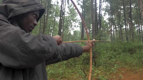 un primer plano orbitando alrededor de la cara de un cazador africano encapuchado mientras apunta con un arco y una flecha tradicionales y dispara mientras caza en un bosque