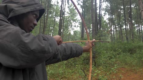 a close up shot orbiting around a hooded african hunters face as he takes aim with a traditional bow and arrow and shoots while hunting in a forest