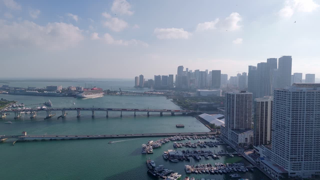 Aerial drone view of downtown Miami skyline with cruise ships docked at the port, scenic Biscayne Bay, and modern urban architecture under a partly cloudy sky.