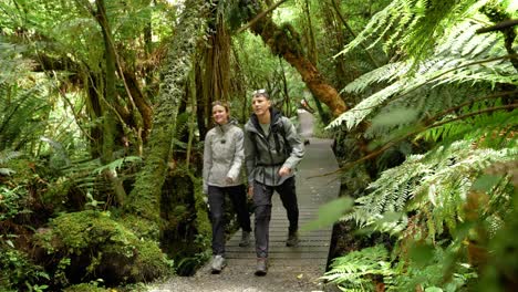 Adventurous-couple-walks-over-foot-bridge-in-mossy-forest-hiking-with-outdoor-gear-in-New-Zealand,-static-full-body