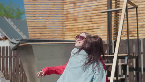 two young women embrace joyfully under clear sky in outdoor setting, one smiling brightly in sunglasses while wrapped in denim, conveying warmth, affection, and friendship
