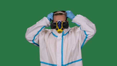 portrait of a male doctor in a white protective suit and surgical gloves looking at the camera puts on a medical respirator and safety glasses. green screen, chroma key. slow motion