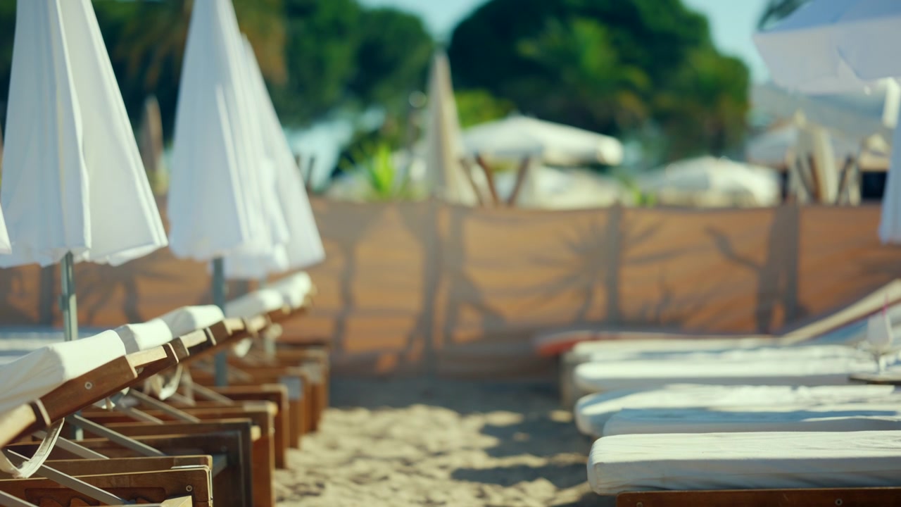 Lettini da spiaggia vuoti disposti su sabbia soffice sotto ombrelloni bianchi.