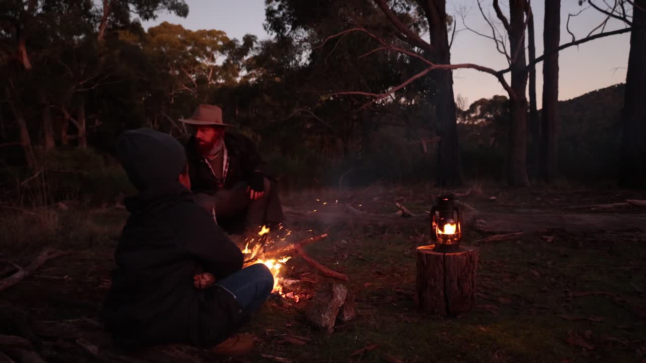 A slow motion shot of a fire crackling out in the Australian bush with a bushman and his son sitting around the fire.