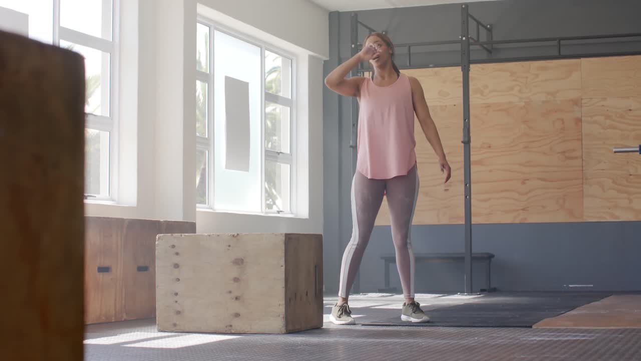cansada mujer biracial inalterada tomando un descanso del entrenamiento cruzado en el gimnasio, cámara lenta