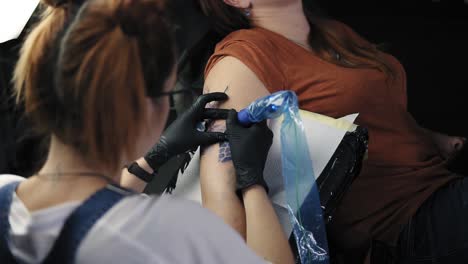 the tattoo artist at work. a red headed girl holding a tattoo machine and applied in a pattern on the skin of her female client. blue ink in the tattoo machine. high angle footage from the shoulder