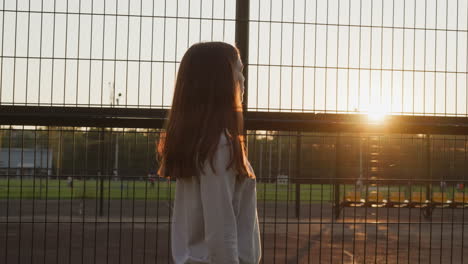 tired lady passes by fence of stadium at sunset. young woman walks after fitness classes looking at football players in evening. feeling of fatigue