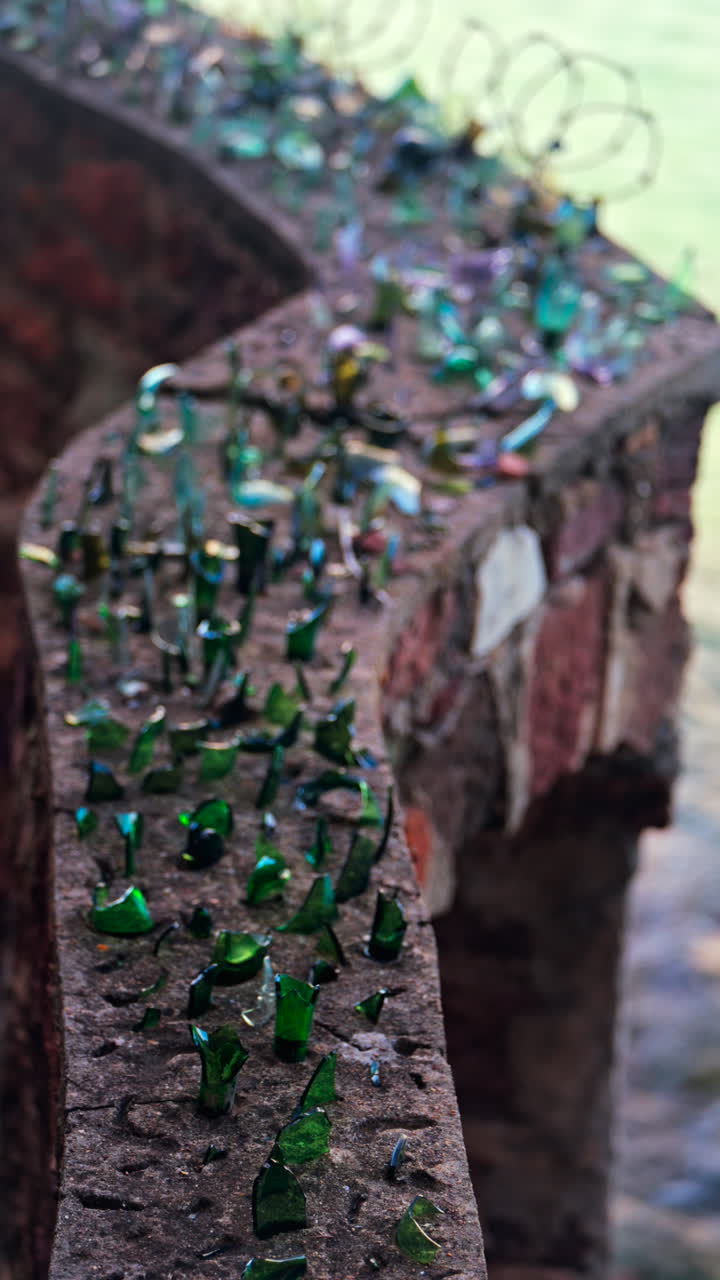 Close up of multiple broken glass shards and barbed wire loops along the edge of a brick wall with a blurred view of the sea. Vertical