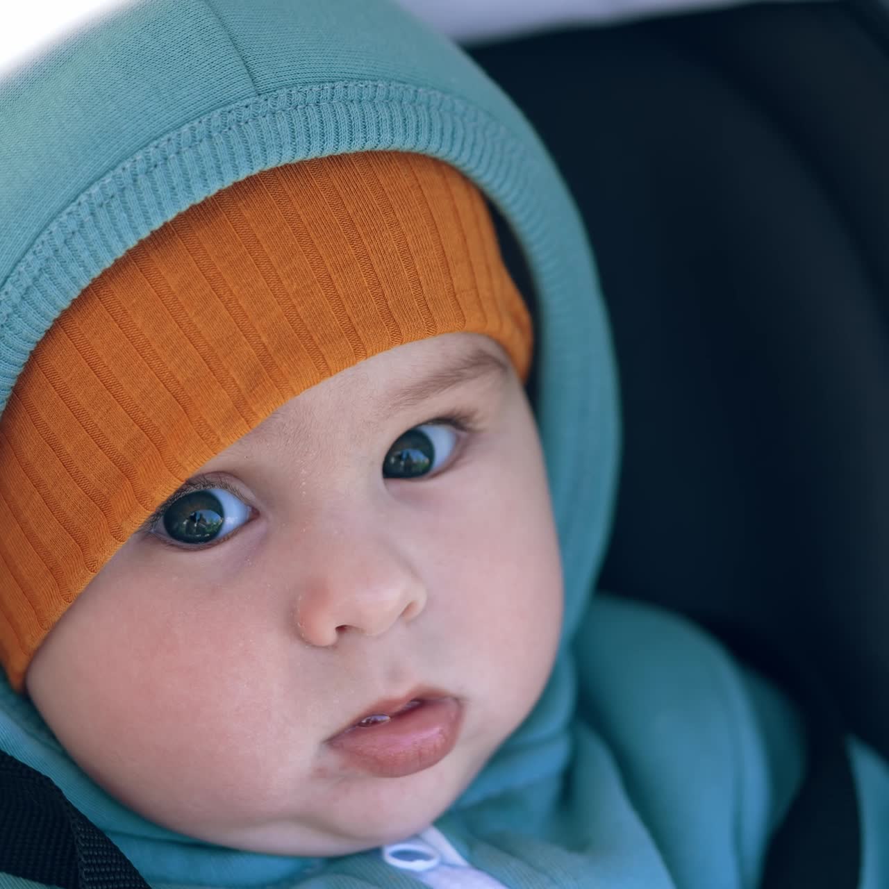 Lovely Caucasian boy with sweet cheeks sits in stroller. Cute child wearing warm clothes and cap having a walk in pram. Close up