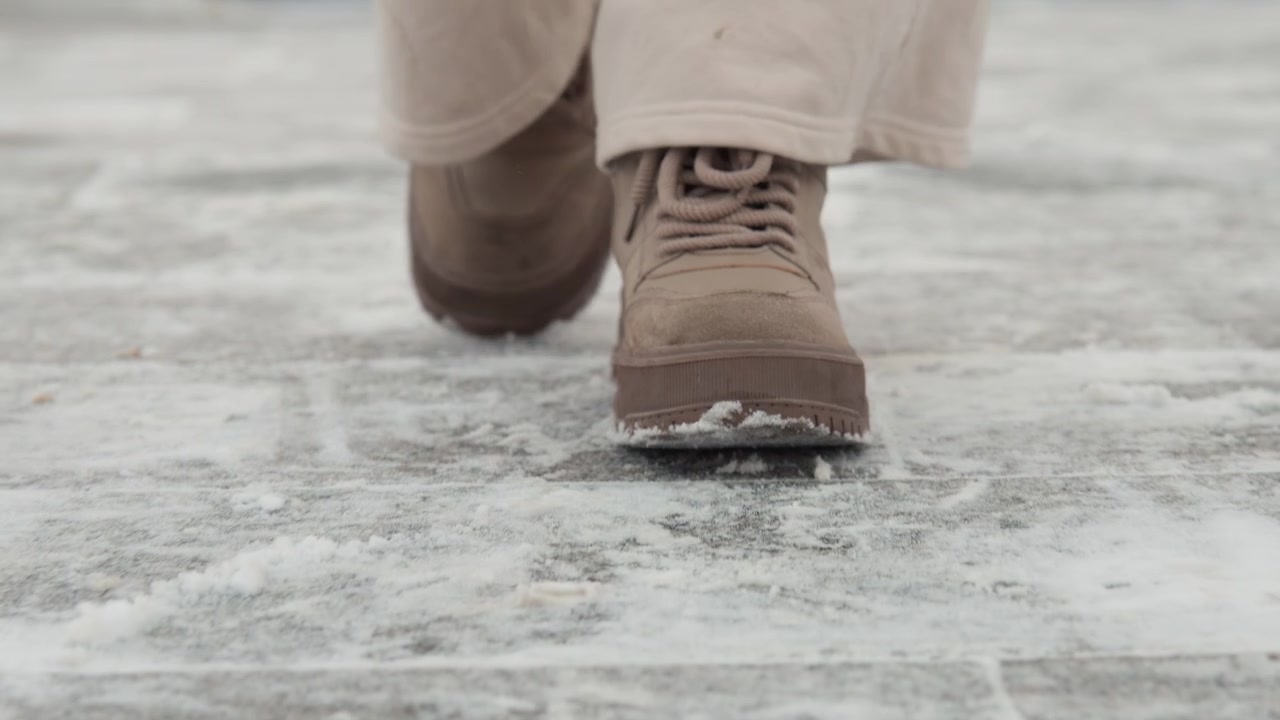 Low-angle close-up of brown winter boots walking across icy, textured pavement with one boot lifted and visible frost scraped along sole; neutral daylight and shallow depth emphasize footwear motion