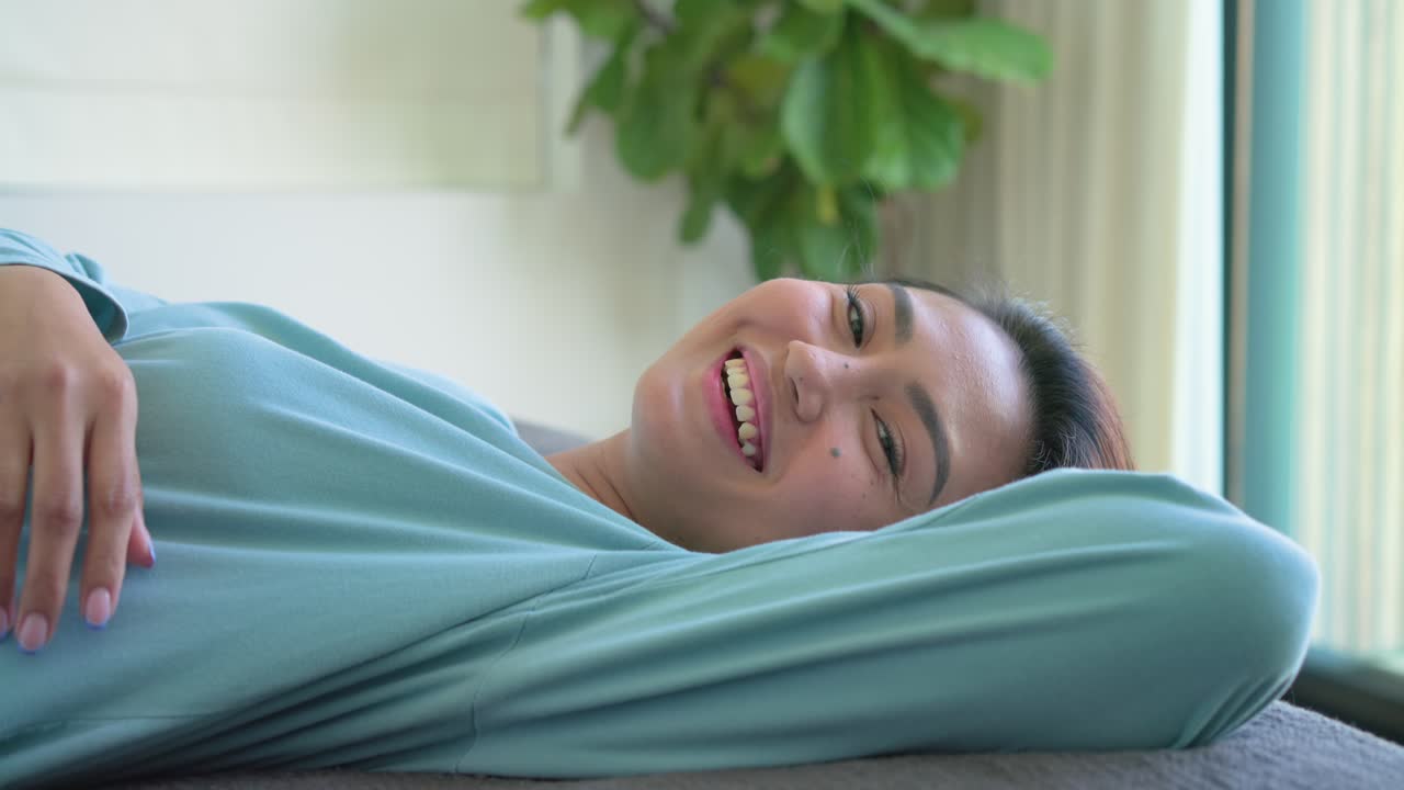 Donna asiatica che apre le palpebre, nota la fotocamera, sorride, sdraiata sul letto vicino alla porta a vetri