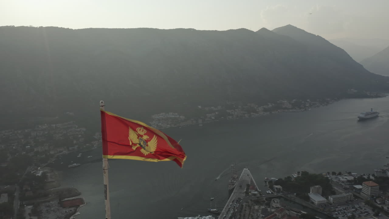 Parallax drone shot of Montenegro's flag waving at dusk with an amazing view of the bay and the mountains