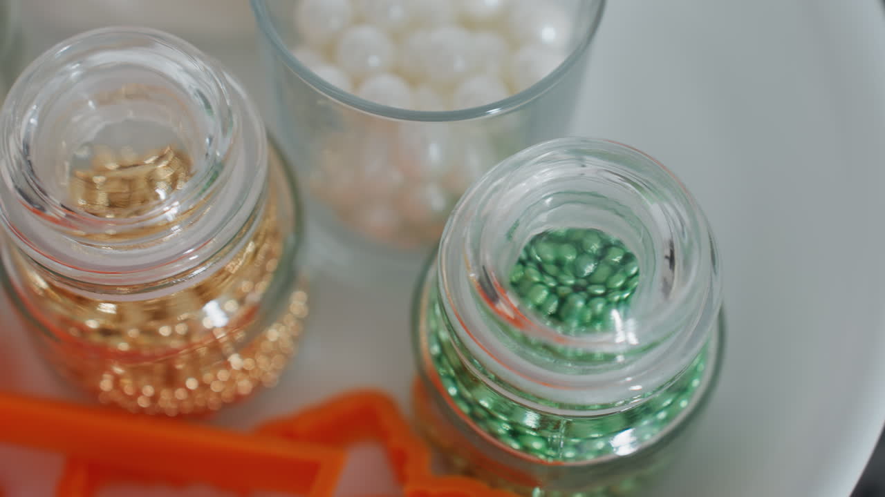 유리 잔에 보관된 활기찬 금과 녹색 장식 구슬과 유리 컵에 있는  진주 구슬, 배경에는 오렌지색 쿠키 절단기, 베이킹을 위해 끔하게 배열되어 있습니다.