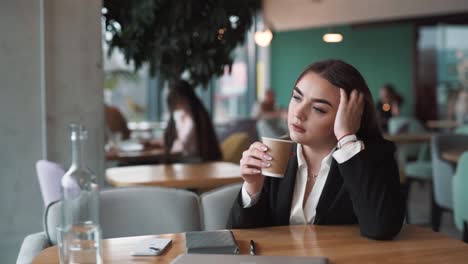 pensive, beautiful woman sits in a stylish cafe in business attire, attentively sipping coffee