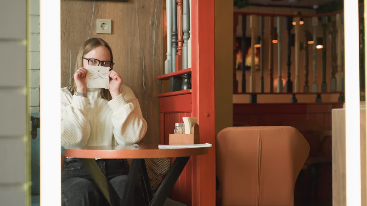 Lady in white sweater and glasses sketching cat mask on napkin at wooden table in cozy cafe, preparing to lift drawing to face while animated flame appears on screen in background