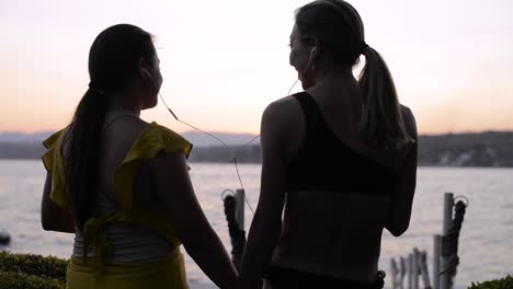 young latin girlfriends sharing earplugs listening to music and dancing near a lake at sunset