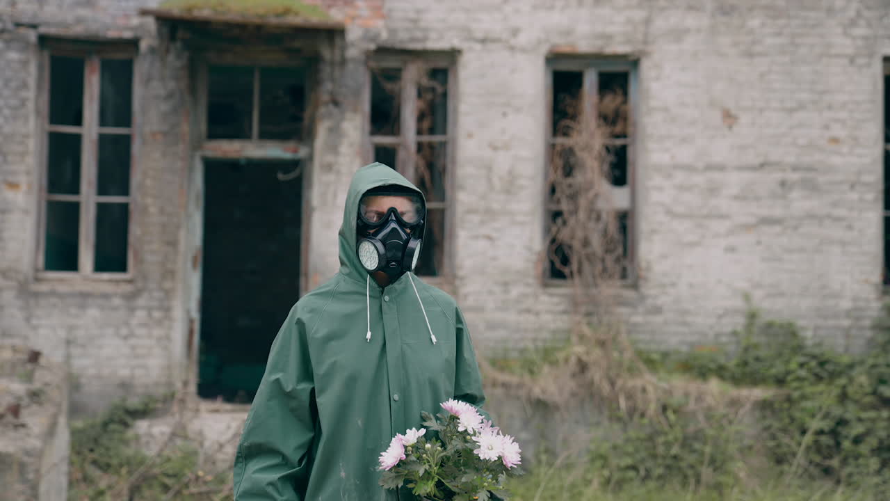 Survivor holding flowers on ruined building background. Person in chemical safety suit and respirator standing with flowers in hands in abandoned place. Chemical attack.