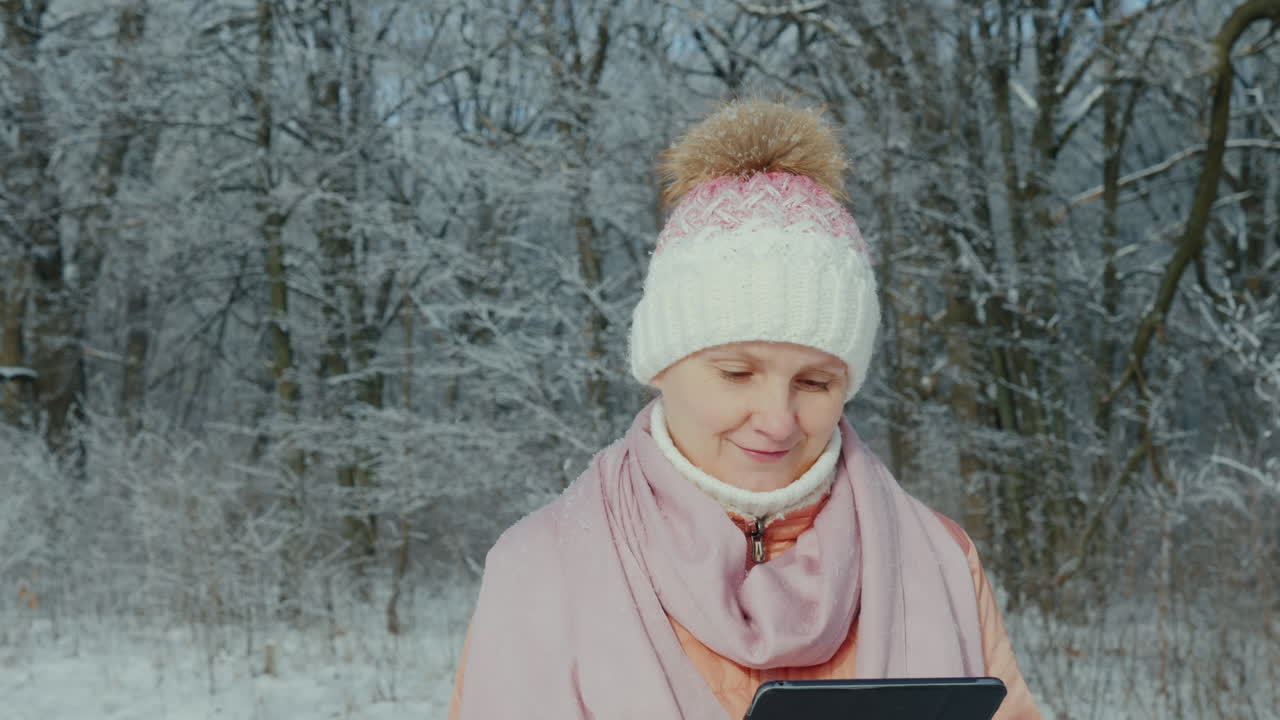 분홍색 재킷을 입은 여성이 디지털 태블릿을 사용하여 겨울 공원에서 산책을 즐깁니다.