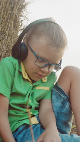 niño pequeño con auriculares profundamente inmerso en la música, sentado en una manta con la cabeza inclinada hacia abajo, rodeado de balas de heno en un entorno rural sereno durante la puesta del sol