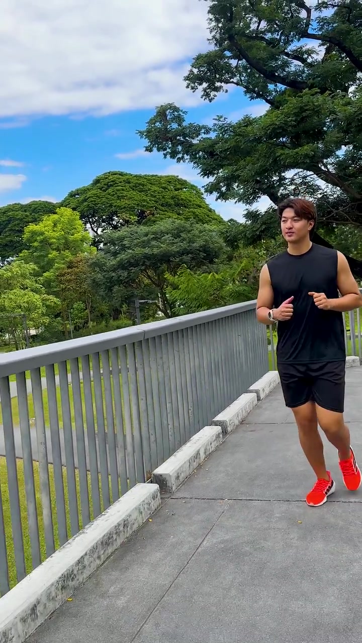 Un uomo asiatico in forma, in abbigliamento sportivo nero, corre verso la telecamera su un ponte in un parco.