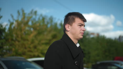 businessman adjusting coat collar while confidently walking outdoors in urban area with background including parked cars, greenery, and office buildings under bright daylight