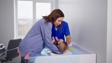 Confident-brunette-girl-puts-her-baby-girl-on-the-bed-in-front-of-an-experienced-male-pediatrician-doctor-in-a-blue-uniform-in-a-pediatric-doctors-office-in-a-modern-clinic