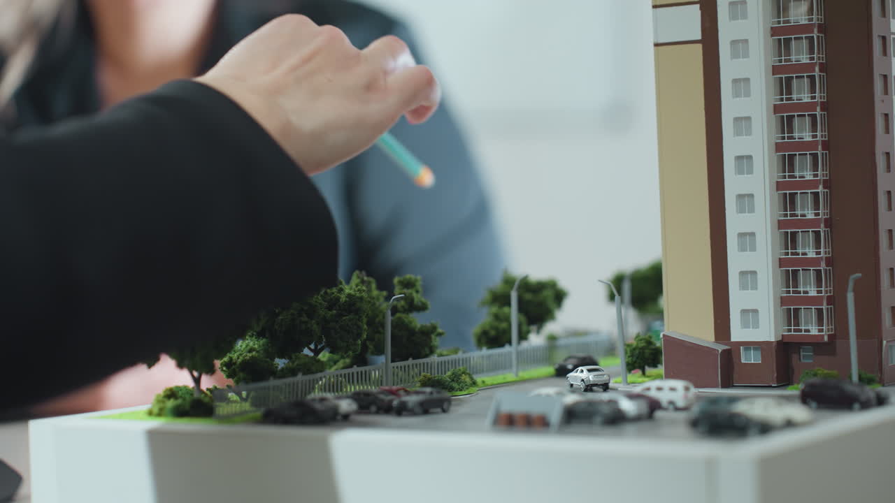 close up hand view of real estate agent holding pencil explaining project to client using architectural building miniature surrounded by small cars trees and road layout in office setting