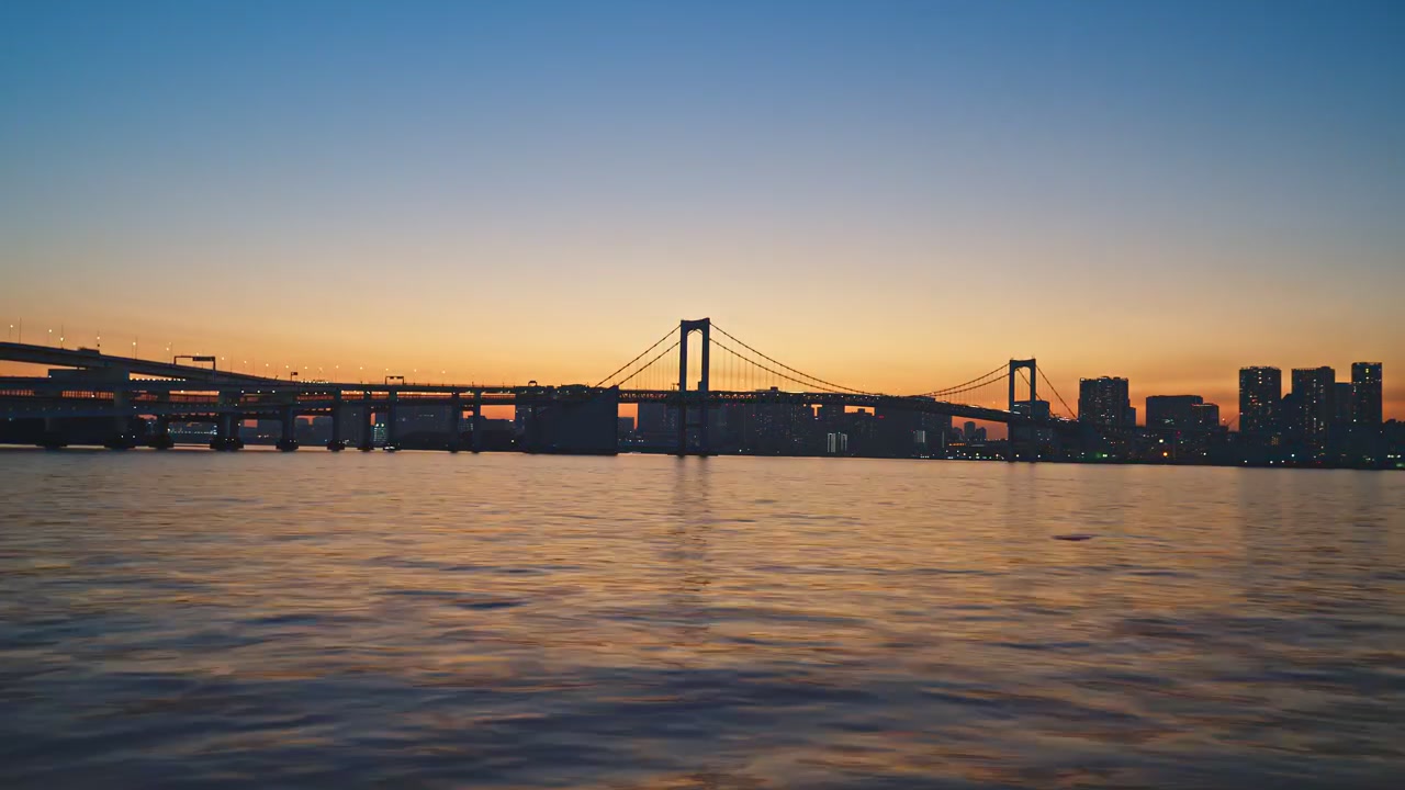 Wide static time-lapse of suspension bridge silhouette over Tokyo Bay, vivid orange sunset band at horizon deepening to rich blue above, calm dark water reflecting warm twilight glow below.